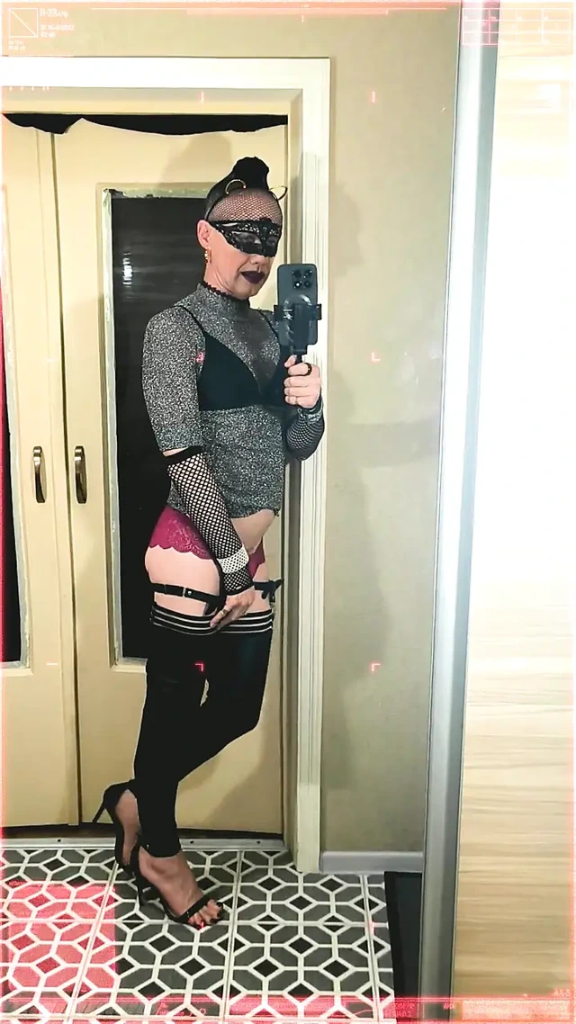 Genç bir kadın kendini aynada kırmızı külot, yüksek topuklu ayakkabılar, maske ve kulaklarla klitorisini okşuyor.