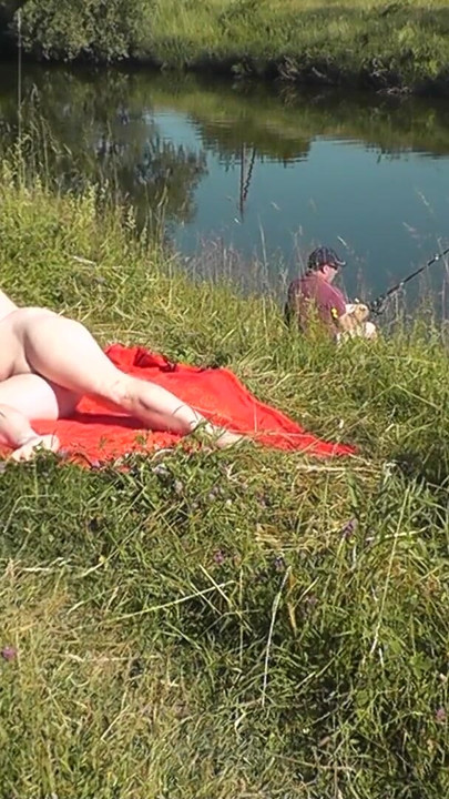 Vahşi plaj. Seksi orta yaşlı seksi kadın platin nehir kıyısında çıplak güneşleniyor, rastgele balıkçı adam izliyor. herkesin içinde çıplak çıplak plaj | Klip 2