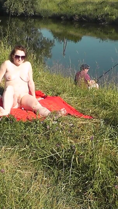 Vahşi plaj. Seksi orta yaşlı seksi kadın platin nehir kıyısında çıplak güneşleniyor, rastgele balıkçı adam izliyor. herkesin içinde çıplak çıplak plaj | Klip 1